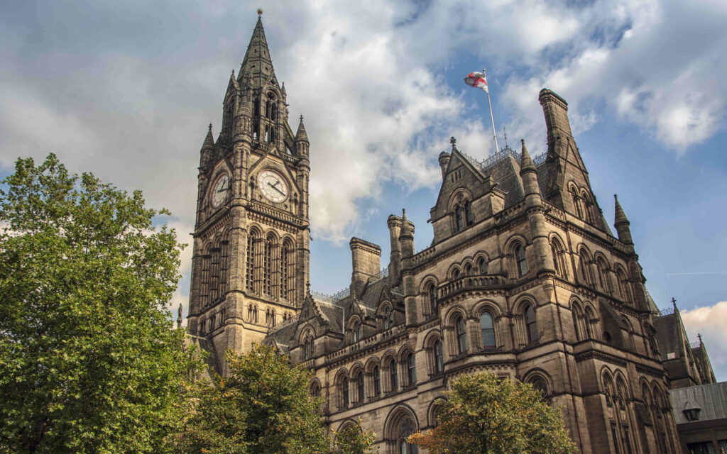 het stadhuis van manchester. het stadhuis heeft een klokkentoren die in dezelfde gotische stijl als de rest van het stadhuis is. om het stadhuis heen zijn diverse bomen te zien.